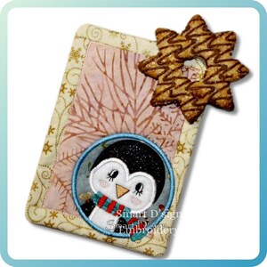 May include: Embroidered Christmas bookmark with a penguin wearing a scarf and mittens. The bookmark has a light pink and beige background, with a star-shaped biscuit attached. The text "Smart D'sign Embroidery" is visible.