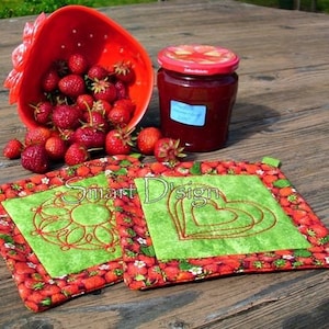 Könnte beinhalten: Zwei grüne und rote gequiltete Topflappen mit gestickten Designs. Ein Topflappen hat ein Herzdesign und der andere ein Blumendesign. Die Topflappen liegen auf einem Holztisch mit einer Schüssel Erdbeeren und einem Glas Erdbeermarmelade.