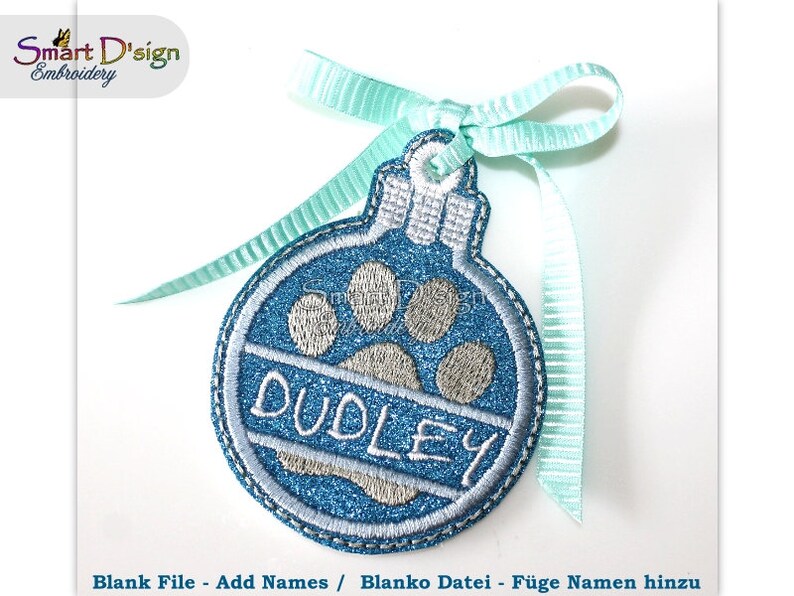 May include: A blue and silver Christmas ornament with a paw print and the name "DUDLEY" embroidered on it. The ornament is tied with a light blue ribbon.
