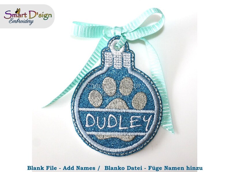 May include: A blue and white Christmas ornament with a paw print and the name "DUDLEY" embroidered on it. The ornament is attached to a light blue ribbon.