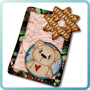 May include: A rectangular, fabric-covered item with a teddy bear design and a star-shaped cookie on top. The fabric has a floral and leaf pattern with a black border. The teddy bear has a red heart and is surrounded by a blue circle.