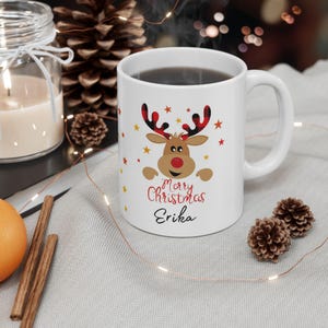 Puede incluir: Taza de cerámica blanca con un diseño de reno navideño. El reno tiene cuernos a cuadros rojos y negros, una nariz roja y estrellas amarillas. La taza dice "Merry Christmas Erika" en escritura roja y negra.