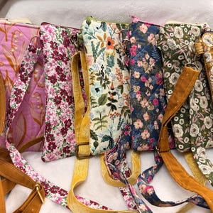 May include: A collection of seven rectangular quilted crossbody bags featuring various floral and botanical patterns in colors including pink, mustard yellow, navy blue, olive green, and white, each equipped with an adjustable fabric shoulder strap.