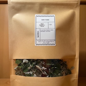 May include: A brown paper bag with a clear window displaying loose black raspberry leaves. The bag has a label that reads "HER THEE" and lists the product as "Black Raspberry Leaves" with a weight of 1 OZ. The label also includes the harvest date.