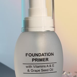 Puede incluir: Botella de cristal esmerilado de prebase de maquillaje con dispensador de bomba transparente. La etiqueta dice "FOUNDATION PRIMER" y enumera las vitaminas A & E y aceite de semilla de uva. Peso neto 28 g.