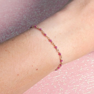 May include: A delicate gold-toned bracelet adorned with small, vibrant pink beads. The bracelet is worn on a person's arm, set against a soft pink background. The beads are evenly spaced along the chain.