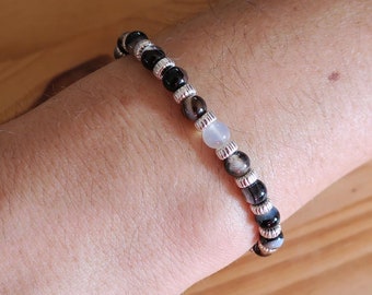 Puede incluir: Un brazalete hecho de cuentas negras, blancas y grises. Las cuentas son redondas y tienen un acabado liso. El brazalete se lleva en la muñeca.
