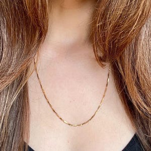 Puede incluir: Un delicado collar de color dorado con un dise&ntilde;o sutil y texturizado. La cadena es visible sobre un fondo oscuro, creando un contraste que resalta la joya. El collar es un accesorio sencillo pero elegante.