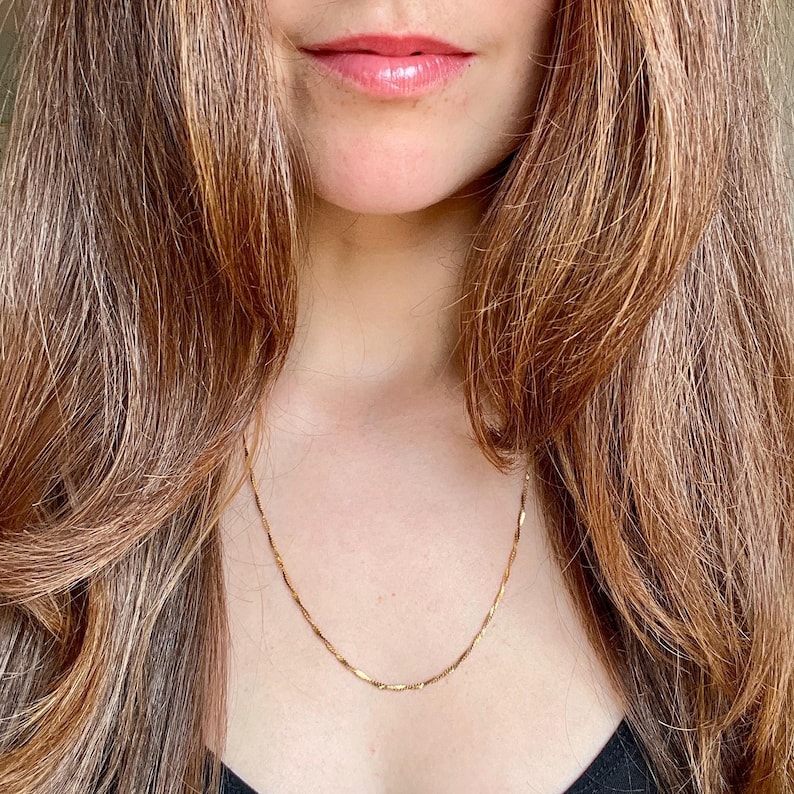Puede incluir: Un delicado collar de color dorado con un dise&ntilde;o sutil y texturizado. La cadena es visible sobre un fondo oscuro, creando un contraste que resalta la joya. El collar es un accesorio sencillo pero elegante.