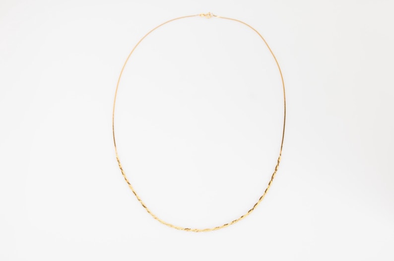 Puede incluir: Un delicado collar de oro con un dise&ntilde;o de cuerda trenzada. El collar se muestra sobre un fondo blanco, mostrando sus finos detalles y su elegante simplicidad. La cadena es delgada y el cierre es visible.