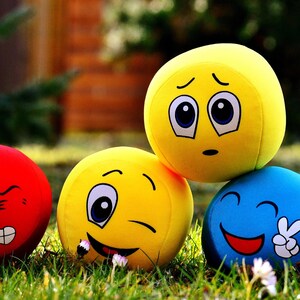 May include: A group of emoji plush balls in red, yellow, and blue, each with a different facial expression. The yellow emoji balls are stacked. The plush toys are set on green grass. The image has a shallow depth of field.