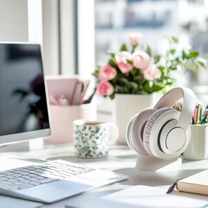 Op de afbeelding: Een lichte werkplek met een laptop, witte koptelefoon en een vaas met roze rozen. Een mok met patroon en kantoorbenodigdheden staan ook op het bureau, alles in natuurlijk licht.
