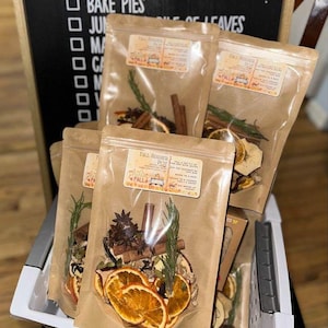 May include: Four brown paper bags filled with dried fruit, spices, and herbs. The bags are labeled "Fall Simmer Pot" and have a small illustration of a house with a chimney. The bags are in a white wicker basket.