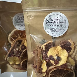 May include: Two clear bags filled with dried fruit and spices, including orange slices, cinnamon sticks, and cranberries. Each bag has a circular label that reads "Winter Wonderland Simmer Pot" with ingredient details. Aromatic potpourri.