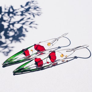 A plant shadow in the left corner, two elongated oval earrings in silver and bright colors: in each one there is an colorful image of red poppies with light green stems and leaves. The colors are vivid and transparent like a stained glass window art.
