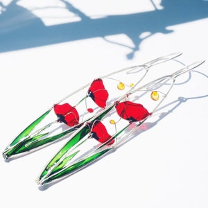 A shadow in the background and two elongated oval earrings in silver and bright colors: in each one there is an colorful image of red poppies with light green stems and leaves. The colors are vivid and transparent like a stained glass window art.