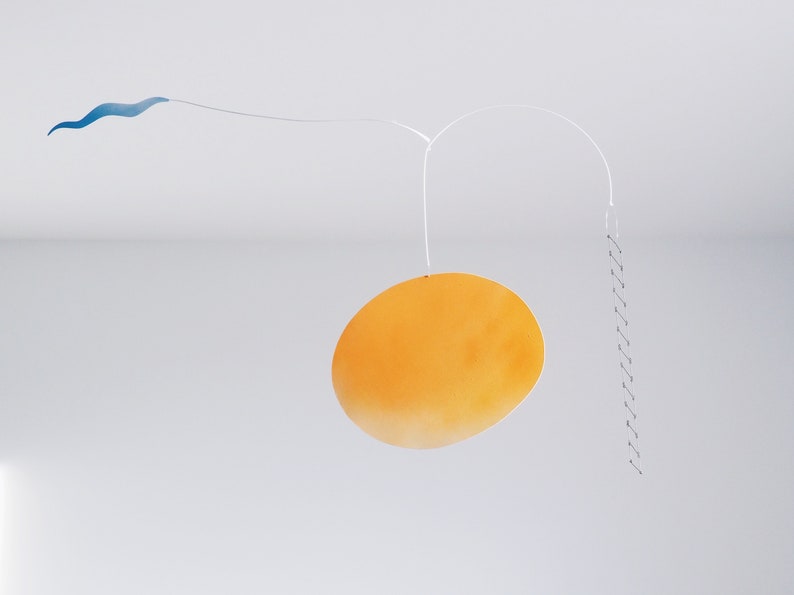 A hanging piece, you can see thin blue cloud in the left, in the middle a vivid orange sun, and a thin black ladder on the right. They are balanced on a thin white wires. There is a white wall and ceiling behind the piece.