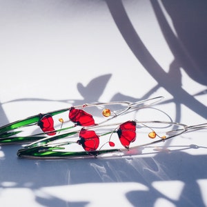 White background and shadows. two long earrings in silver and bright colors: in each one there is an colorful picture created with silver wire:red poppies with light green stems. The colors are vivid and transparent like a stained glass window art.