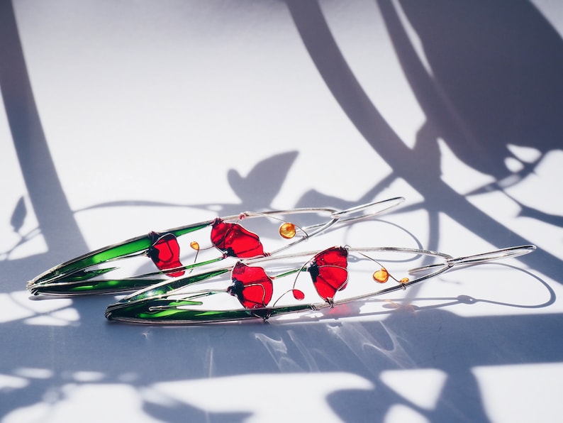 White background and shadows. two long earrings in silver and bright colors: in each one there is an colorful picture created with silver wire:red poppies with light green stems. The colors are vivid and transparent like a stained glass window art.