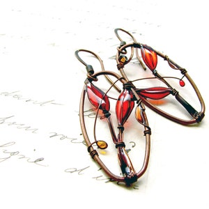 On white sheet with elegant handwriting lie two earrings in dark brown copper and bright colors - red and  yellow. The colors are transparent, colorful shadows can be seen on the paper. In the ovals we can see an image of red plant created with wire