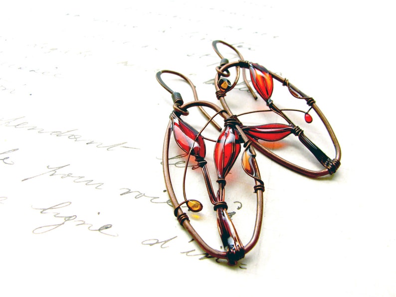 On white sheet with elegant handwriting lie two earrings in dark brown copper and bright colors - red and  yellow. The colors are transparent, colorful shadows can be seen on the paper. In the ovals we can see an image of red plant created with wire