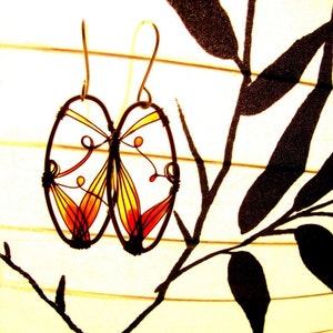The two dark oval earrings are hanging on a paper lamp with a black bamboo drawing. We can see that each earring has a design made with thin wire and colored in yellow to dark amber colors. The colors are transparent and vivid.