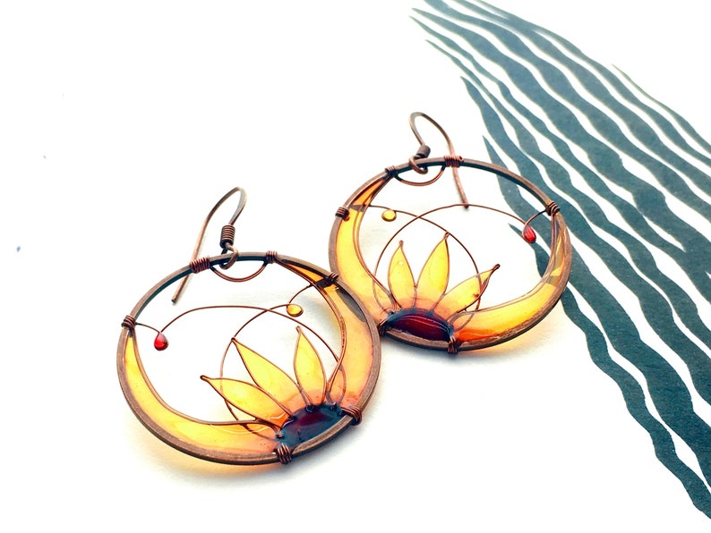 Two round earrings in dark copper wire with a half sunflower in each one, colored in yellow, amber and brown.