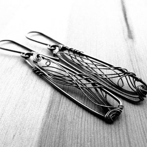 Black and white photo. In the center two dark grey earrings with a triangle shape are laying on a wooden surface. In each earring there is an intricate design created with thin wire.