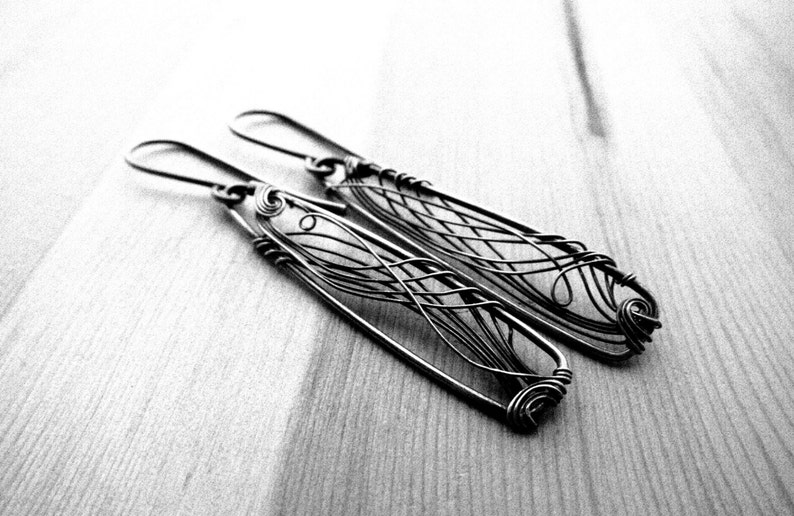 Black and white photo. In the center two dark grey earrings with a triangle shape are laying on a wooden surface. In each earring there is an intricate design created with thin wire.