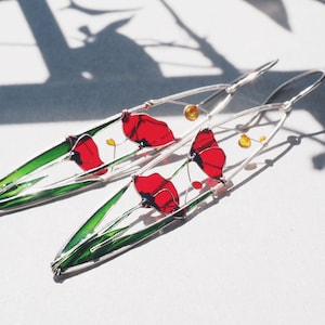 A shadow in the background and two elongated oval earrings in silver and bright colors: in each one there is an colorful image of red poppies with light green stems and leaves. The colors are vivid and transparent and leave a colorful shadow.