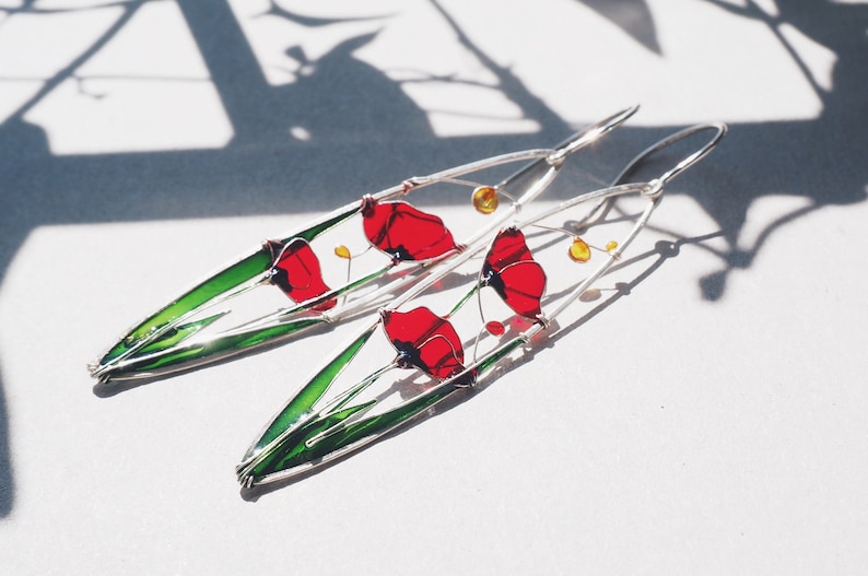 A shadow in the background and two elongated oval earrings in silver and bright colors: in each one there is an colorful image of red poppies with light green stems and leaves. The colors are vivid and transparent and leave a colorful shadow.