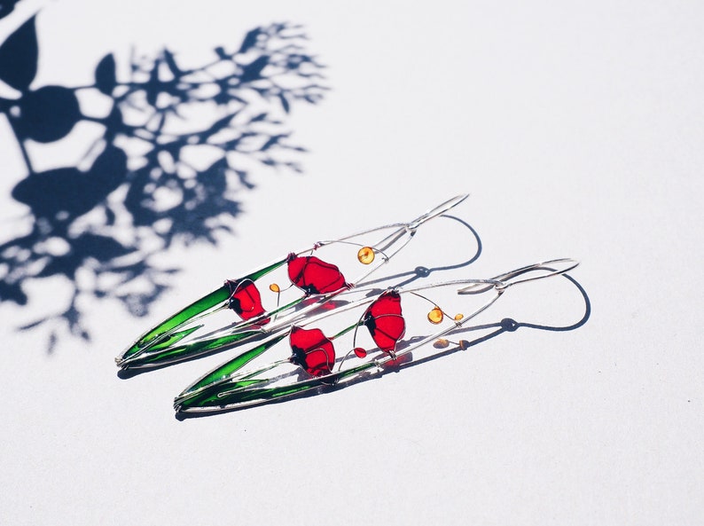 A plant shadow in the left corner, two elongated oval earrings in silver and bright colors: in each one there is an colorful image of red poppies with light green stems and leaves. The colors are vivid and transparent like a stained glass window art.