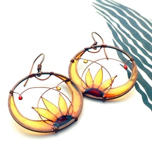 Two circle-shaped earrings in dark copper, each earring contains a half sunflower made with wire and colored in yellow, amber and brown transparent colors.