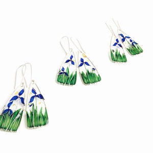 On white background there are three pairs of earrings. Each one is slightly different but features blue iris flowers, light green stems and grass and a touch of yellow sunshine.