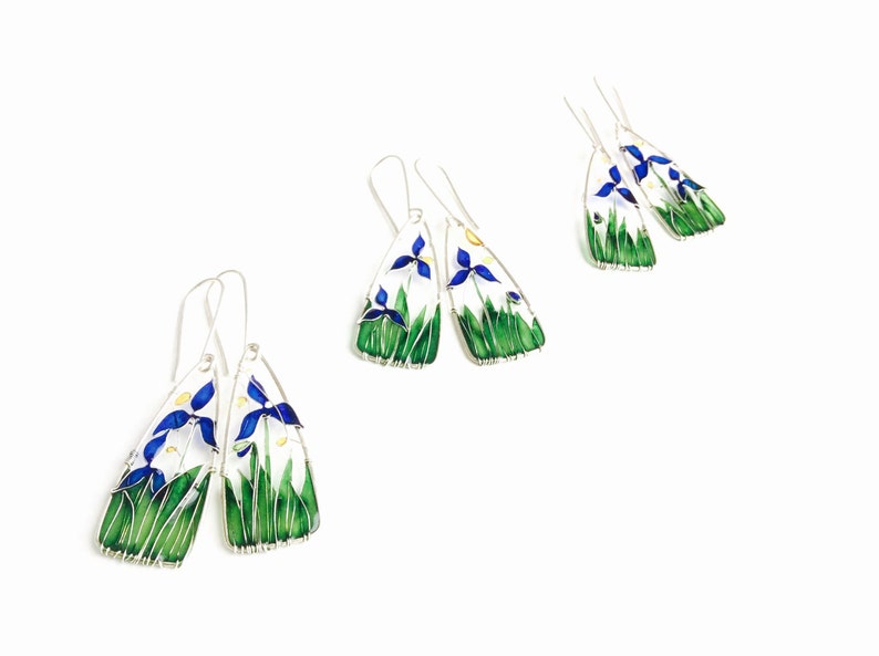 On white background there are three pairs of earrings. Each one is slightly different but features blue iris flowers, light green stems and grass and a touch of yellow sunshine.