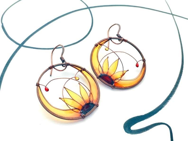 Two circle-shaped earrings in dark copper, each earring contains a half sunflower made with wire and colored in yellow, amber and brown transparent colors.