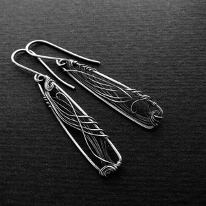 Black background, a dark photo. In the center, situated diagonally, there are two earrings with in a long triangle shape. In each triangle there are elegant lines created with thin wire.