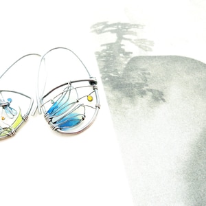 Two earrings in dark silver color on a book page. There is a little colorful landscape created with wire for each earring. The two pictures are mismatched but with the same colors: Light green, vivid yellow and light to darker blue for the dragonfly.