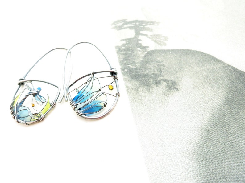 Two earrings in dark silver color on a book page. There is a little colorful landscape created with wire for each earring. The two pictures are mismatched but with the same colors: Light green, vivid yellow and light to darker blue for the dragonfly.