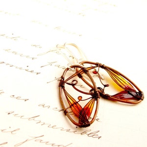 On a handwritten notebook page lies a pair of earrings. They have oval shape made with dark brown metal wire and each earring has a colorful detail in amber to light yellow colors. The colors are transparent, the ear wires are silver.