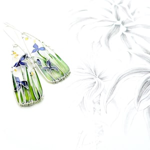 On white background with light grey sketches lies a pair of earrings. They have triangle shapes and each includes a landscape of iris flowers, green grass, and a touch of sunshine, all made with thin silver wire. The two pictures are mismatched.