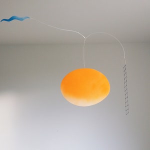 A hanging piece, you can see thin blue cloud in the left, in the middle a vivid orange sun, and a thin black ladder on the right. They are balanced on a thin white wires. There is a grey wall behind the piece.