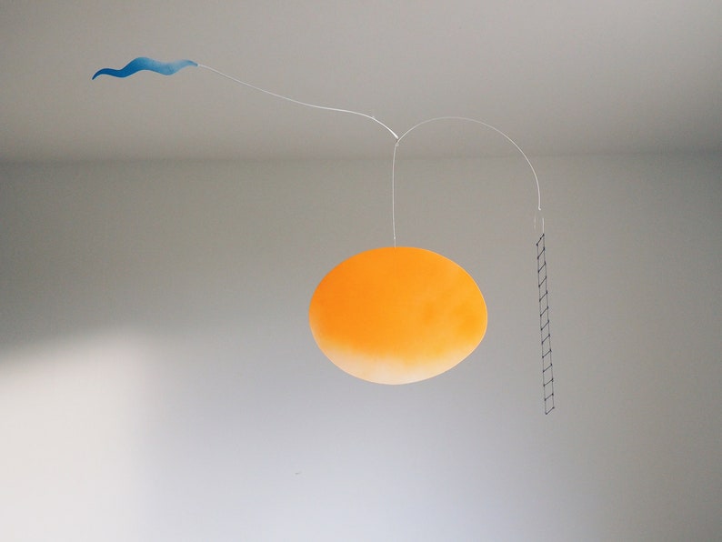 A hanging piece, you can see thin blue cloud in the left, in the middle a vivid orange sun, and a thin black ladder on the right. They are balanced on a thin white wires. There is a grey wall behind the piece.