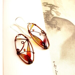 On a page with black and white illustration lie two amber colored earrings. They have oval shape made with dark brown metal wire and each earring has a colorful detail in amber to light yellow. The colors are transparent, the ear wires are silver.