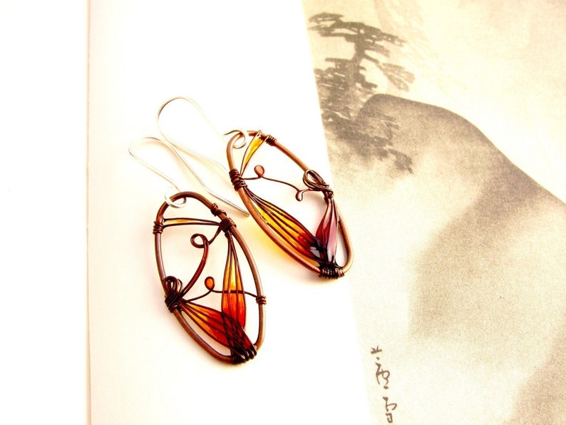 On a page with black and white illustration lie two amber colored earrings. They have oval shape made with dark brown metal wire and each earring has a colorful detail in amber to light yellow. The colors are transparent, the ear wires are silver.