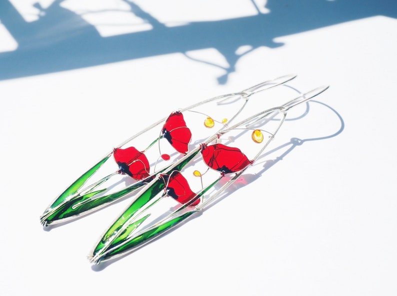 A shadow in the background and two elongated oval earrings in silver and bright colors: in each one there is an colorful image of red poppies with light green stems and leaves. The colors are vivid and transparent like a stained glass window art.