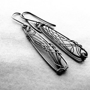 Black and white photo. In the center there are two long, dark grey earrings with a triangle shape. In each earring there is an intricate design created with thin wire.