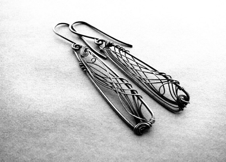 Black and white photo. In the center there are two long, dark grey earrings with a triangle shape. In each earring there is an intricate design created with thin wire.