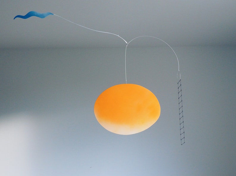 A hanging piece, you can see thin blue cloud in the left, in the middle a vivid orange sun, and a thin black ladder on the right. They are balanced on a thin white wires. There is a grey wall behind the piece.