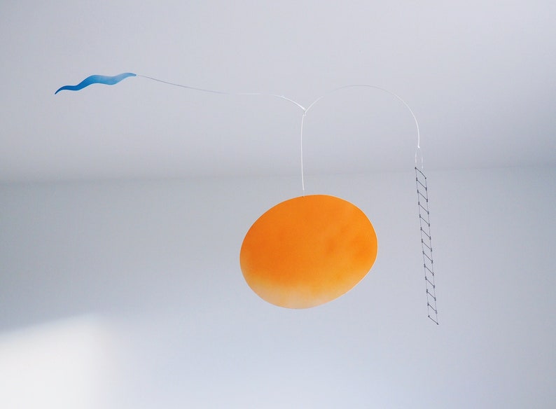 A hanging piece, you can see thin blue cloud in the left, in the middle a vivid orange sun, and a thin black ladder on the right. They are balanced on a thin white wires. There is a white wall behind the piece.